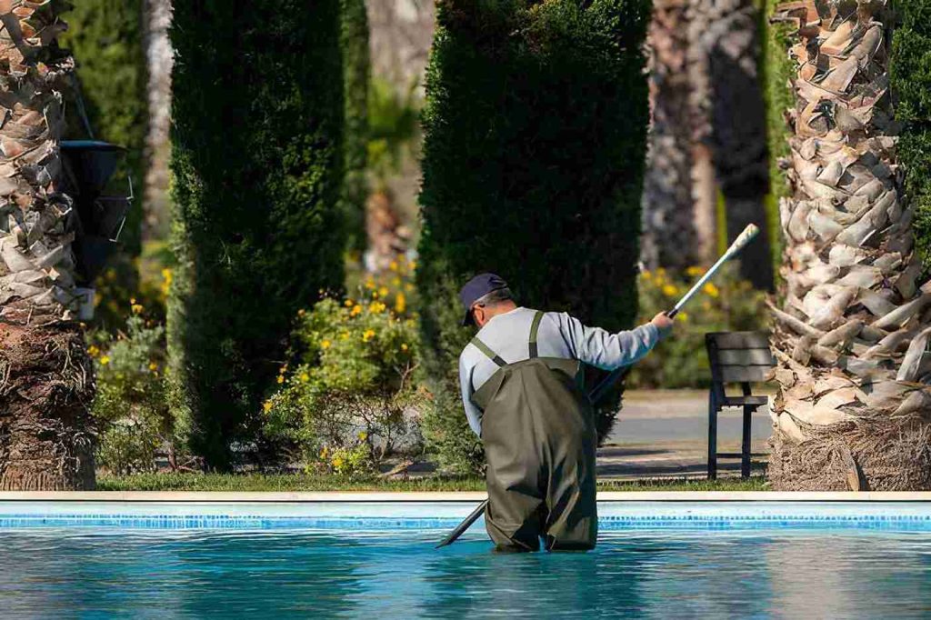 Hobe Sound pool service technician wearing waders cleaning a tropical outdoor swimming pool with a net pole.
