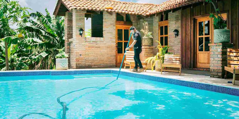 Cleaning pool service Jensen Beach professional using long pole to clean clear blue pool beside rustic brick house.