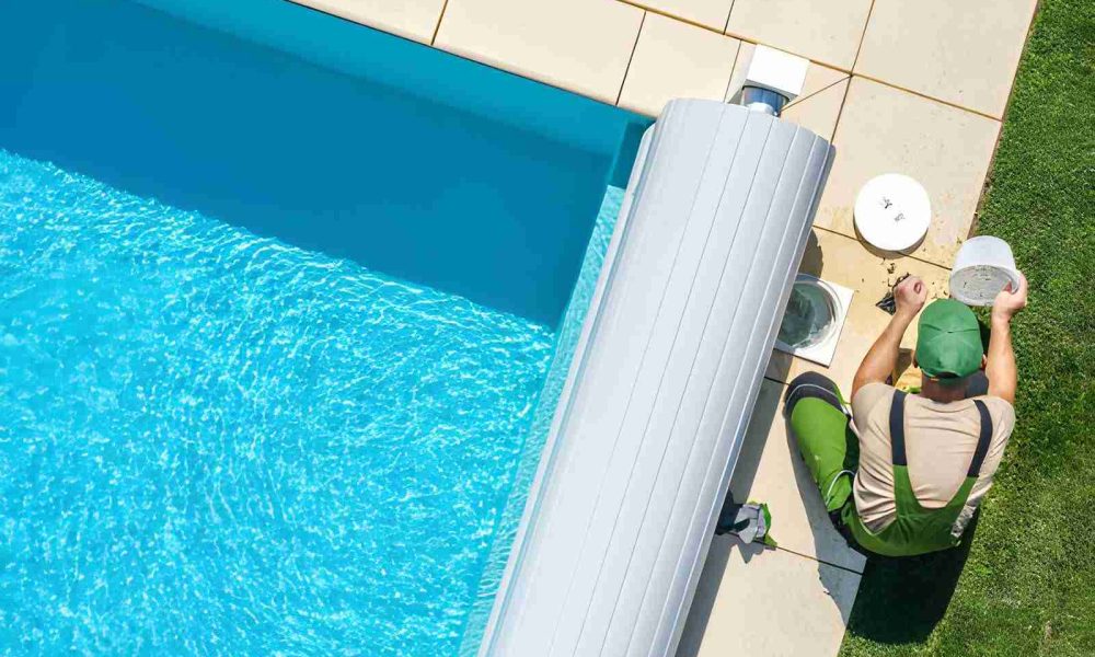 Pool maintenance Port St Lucie technician in green overalls servicing filtration system beside clear blue swimming pool.