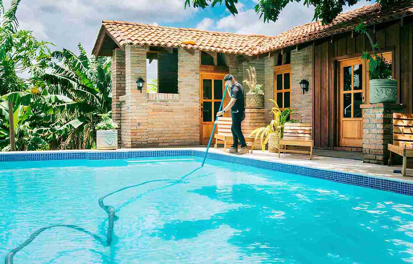 Cleaning pool service Jensen Beach professional using long pole to clean clear blue pool beside rustic brick house.