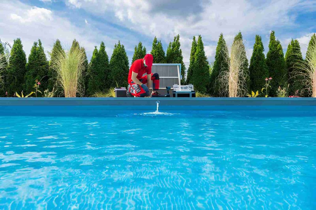 Pool maintenance Port St Lucie technician in red uniform inspecting and cleaning clear blue swimming pool outdoors.