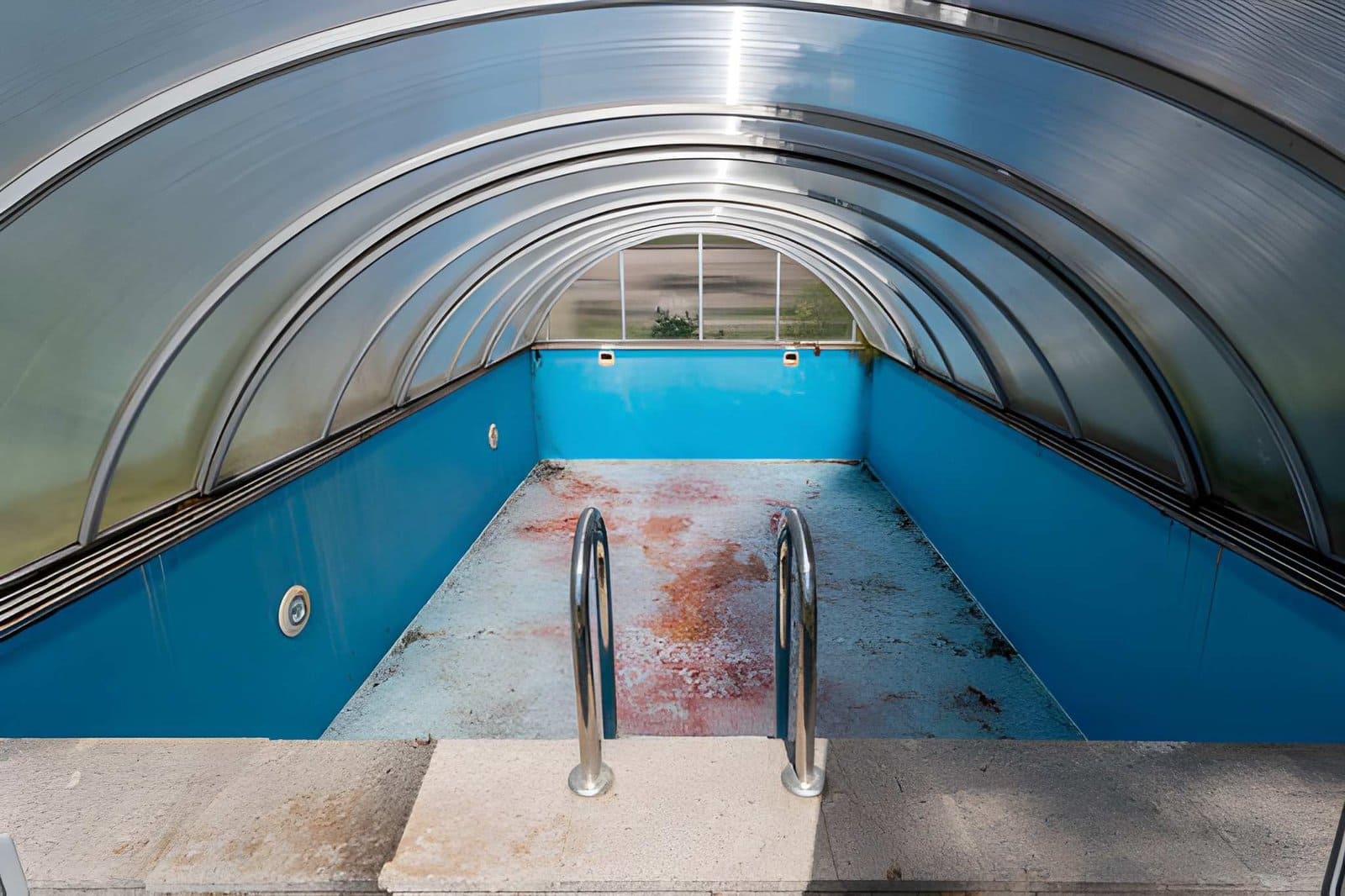 Pool enclosure repair Jensen Beach FL empty covered swimming pool with blue interior and metal handrails.
